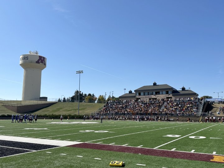 A lively crowd of students, decked out in maroon and gold, cheers enthusiastically from the stands during a game at the University of Minnesota Morris. Many fans raise their hands in excitement, waving and showing their school spirit. The iconic water tower with the University of Minnesota 'M' logo looms in the background